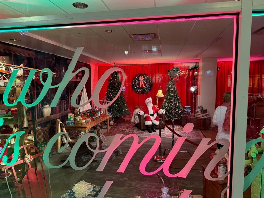 View through the storefront window lettering reading 'guess who is coming to town' with Santa seated inside